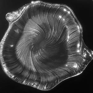 May include: Clear glass decorative bowl with a swirling pattern. The bowl has an irregular, wavy edge and a spiral design radiating from the center. The glass reflects light, creating highlights against the dark background.