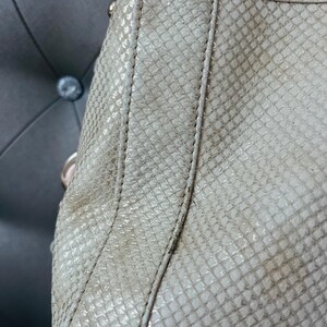 Pu&ograve; includere: Primo piano di una borsa beige con motivo a pelle di serpente. La borsa ha un design strutturato con cuciture visibili lungo le cuciture. La trama appare leggermente usurata, con sottili variazioni di colore.