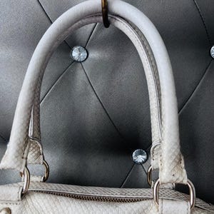 Pu&ograve; includere: Borsa a mano bianco sporco con motivo a pelle di serpente, con manico superiore e chiusura a zip. La borsa ha finiture color argento ed &egrave; appesa a un gancio in ottone su uno sfondo grigio a forma di diamante.