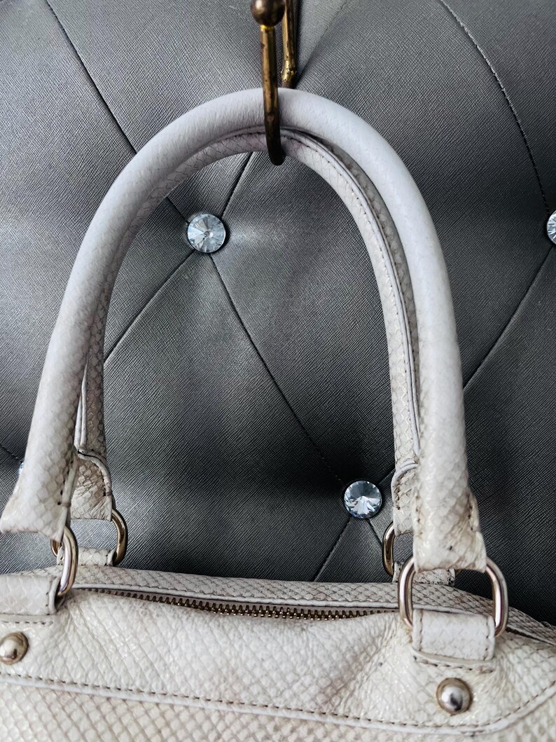 Pu&ograve; includere: Borsa a mano bianco sporco con motivo a pelle di serpente, con manico superiore e chiusura a zip. La borsa ha finiture color argento ed &egrave; appesa a un gancio in ottone su uno sfondo grigio a forma di diamante.