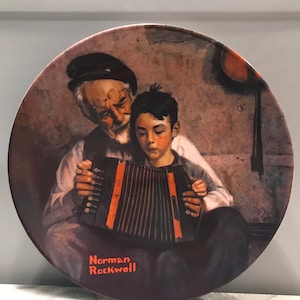 May include: A vintage ceramic plate featuring a painting by Norman Rockwell. The painting depicts an older man with a white beard and a young boy playing an accordion. The plate has a brown background with a rustic feel.
