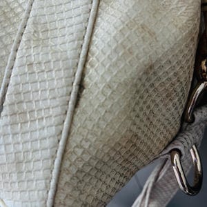 Pu&ograve; includere: Primo piano di una borsa color crema con un motivo strutturato simile a quello di un rettile. La borsa presenta un cinturino bianco con cuciture e finiture metalliche. La superficie della borsa mostra segni di usura.
