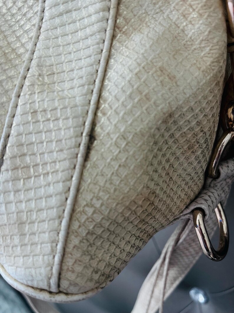 Pu&ograve; includere: Primo piano di una borsa color crema con un motivo strutturato simile a quello di un rettile. La borsa presenta un cinturino bianco con cuciture e finiture metalliche. La superficie della borsa mostra segni di usura.