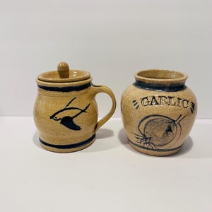 May include: A tan speckled ceramic jar with a lid and a handle, featuring a dark blue painted bird design, alongside a matching tan speckled ceramic garlic keeper with a dark blue painted garlic bulb illustration and the word GARLIC.