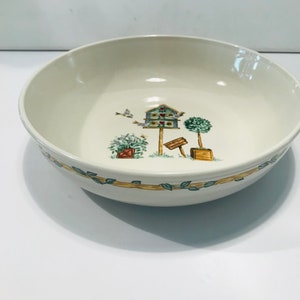 May include: A white ceramic bowl with a green leafy border and a whimsical design of a birdhouse with birds, potted plants, and a sign.