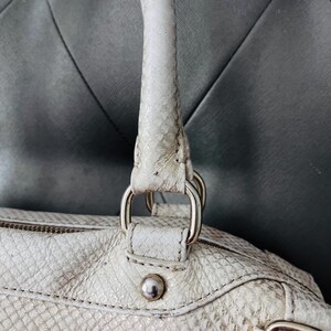 Pu&ograve; includere: Borsa a mano color crema con un motivo strutturato simile alla pelle di serpente. La borsa presenta un manico superiore, anelli in metallo argentato e un borchia decorativa argentata. Il design della borsa include una chiusura con cerniera e dettagli cuciti.