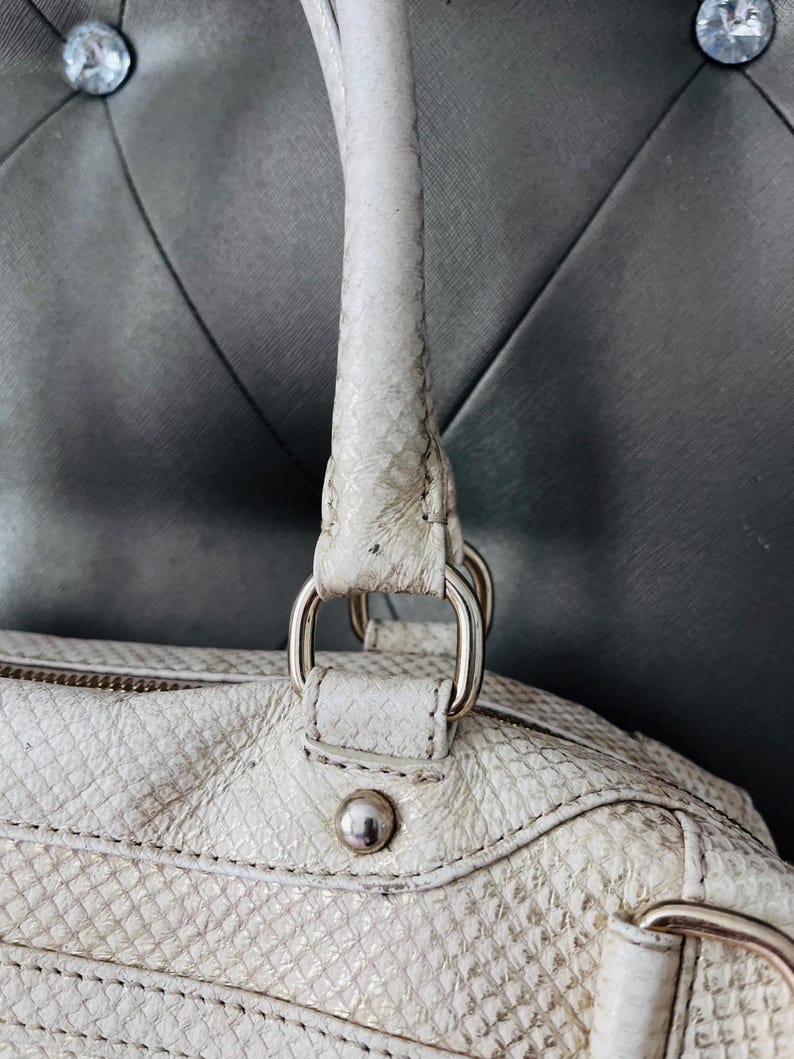 Pu&ograve; includere: Borsa a mano color crema con un motivo strutturato simile alla pelle di serpente. La borsa presenta un manico superiore, anelli in metallo argentato e un borchia decorativa argentata. Il design della borsa include una chiusura con cerniera e dettagli cuciti.