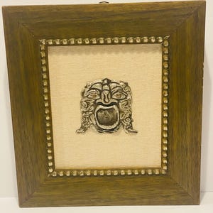 May include: A framed, silver-colored mask with an open mouth, set against a beige fabric background. The mask is framed by a wooden frame with a beaded inner border. The frame is a warm, medium brown color.