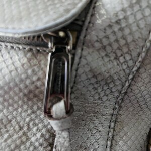 Pu&ograve; includere: Primo piano di una borsa grigio chiaro con un motivo a pelle di rettile. La cerniera lampo &egrave; argentata e ha un piccolo nappa in pelle abbinato. La trama della borsa &egrave; visibile, con dettagli di cucitura.