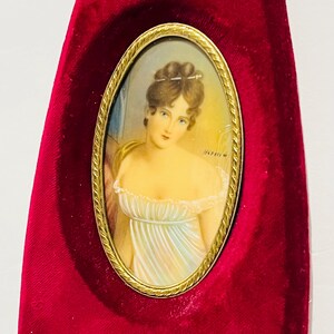 May include: An oval portrait of a woman in a white dress, set in a gold frame, against a deep red velvet background. The portrait features a woman with light skin, brown hair, and blue eyes. The frame is ornate, and the velvet is a rich, saturated color.
