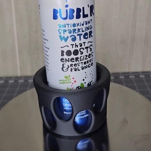 Könnte beinhalten: Eine weiße Dose Bubbl'r Antioxidant Sparkling Water in einem schwarzen Becherhalter. Die Dose trägt den blauen Text "antioxidant sparkling water" und "that boosts, energizes, restores balance". Der Becherhalter hat kreisförmige Aussparungen.
