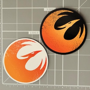 May include: Two circular stickers featuring a stylized bird emblem. One sticker has a white bird on an orange gradient background, while the other has a black bird on an orange gradient background. The stickers are placed on a grid mat with 2.5x3.5 and 3.5x5 inch labels.