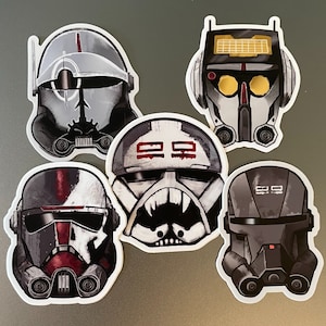 May include: Five Star Wars themed helmet stickers. The helmets are in various shades of gray and black with red accents. The helmets are all different designs and have various details like stripes and markings.
