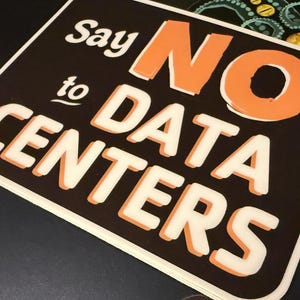 May include: A rectangular sign with a dark brown background and rounded corners. The text reads "Say NO to DATA CENTERS" in a bold, layered font. The word "NO" is in orange, while the other words are in white with orange drop shadows.