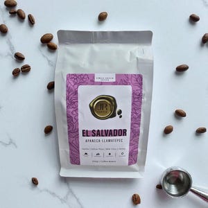 Op de afbeelding: Een witte koffiezak met een paars etiket met de tekst "EL SALVADOR" en "JR COFFEE". Koffiebonen en een zilveren schep omringen de zak. Het etiket geeft de herkomst en de smaaknotities van de koffie aan. Bevat 250g bonen.