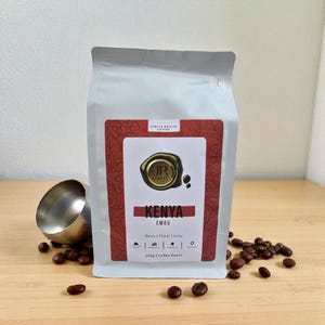 May include: A white bag of Kenya Embu single-origin coffee beans. The bag has a red and white label with the text "JR Coffee" and "Kenya Embu". The bag is on a wooden surface with coffee beans and a metal scoop.