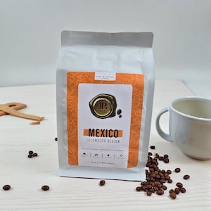May include: A white bag of "JR Coffee" from the Mexico Soconusco Region, with a label featuring the text "Mexico" and "250g Coffee Beans". Coffee beans and a white mug are next to the bag. The label also lists flavour notes.