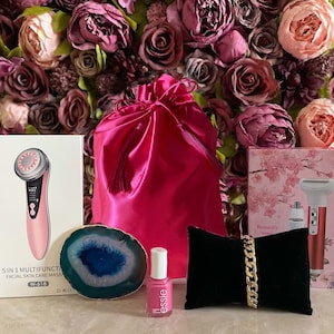 Puede incluir: Un masajeador facial de cuidado de la piel rosa con una pantalla de visualización, una bolsa de satén rosa, una rodaja de ágata dorada, una botella de esmalte de uñas rosa, un brazalete de cadena dorada y un expositor de joyas de terciopelo negro. Los productos están dispuestos sobre un fondo de flores artificiales rosas y moradas.