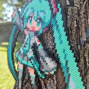 May include: A pixel art figure of a character with long, turquoise pigtails, a gray and white outfit, and pink accents. The figure is made of small, colorful beads and is mounted on a tree trunk.