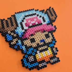 May include: A pixel art creation of a cartoon character with a blue hat, pink tongue, and black and tan features. The character is made of small, colorful beads and set against an orange background.