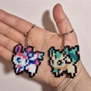 May include: Two pixel art keychains featuring characters from a popular video game. The keychain on the left is pink, blue, and white, while the keychain on the right is beige, green, and white.