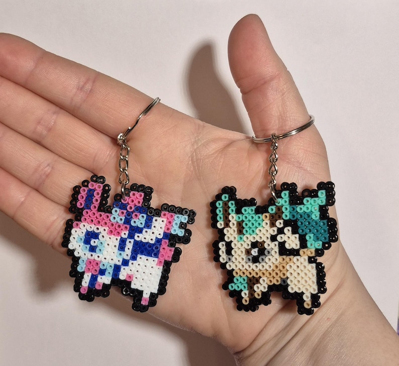 May include: Two pixel art keychains featuring characters from a popular video game. The keychain on the left is pink, blue, and white, while the keychain on the right is beige, green, and white.
