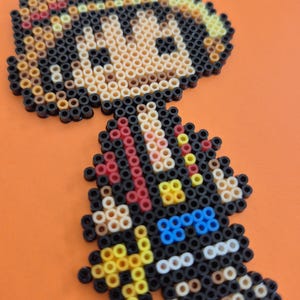May include: A pixel art figure made from colorful plastic beads. The figure has a straw hat with a red band, black hair, and a black outfit with blue and yellow accents. The background is a solid orange.