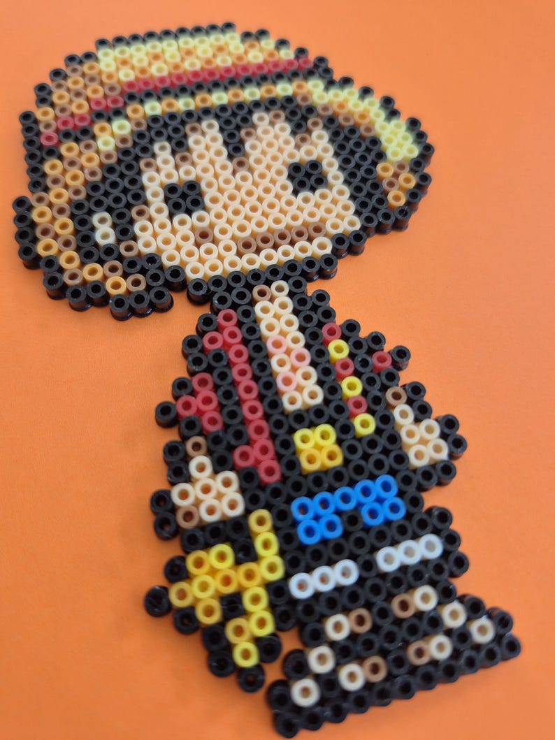 May include: A pixel art figure made from colorful plastic beads. The figure has a straw hat with a red band, black hair, and a black outfit with blue and yellow accents. The background is a solid orange.