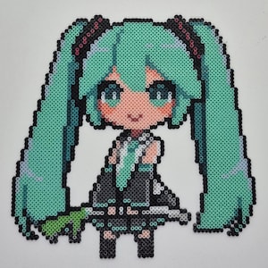 May include: A pixel art depiction of a character with long, turquoise pigtails, large eyes, and a black and gray outfit. The artwork is created using small, colorful beads, giving it a textured appearance. The character is smiling.
