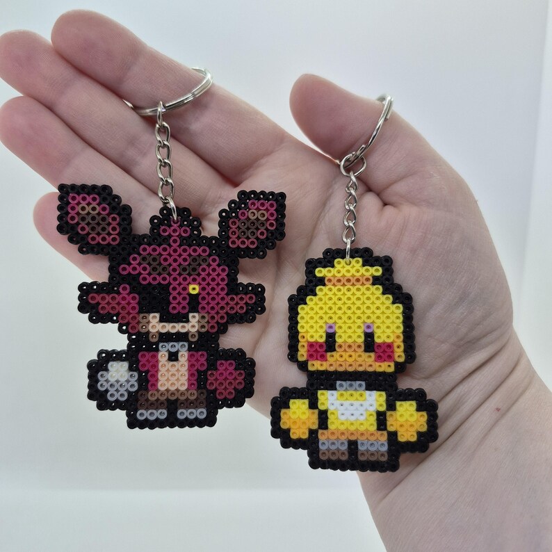 May include: Two pixel art keychains. One is a fox with pink, black, and brown beads. The other is a yellow chick with black and red beads. Both have silver key rings and chains.