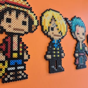 May include: Pixel art of four characters from the anime series One Piece. The characters are made from colorful beads and are arranged on an orange background. The first character is wearing a straw hat. The other characters are Sanji, Zoro, and Chopper.