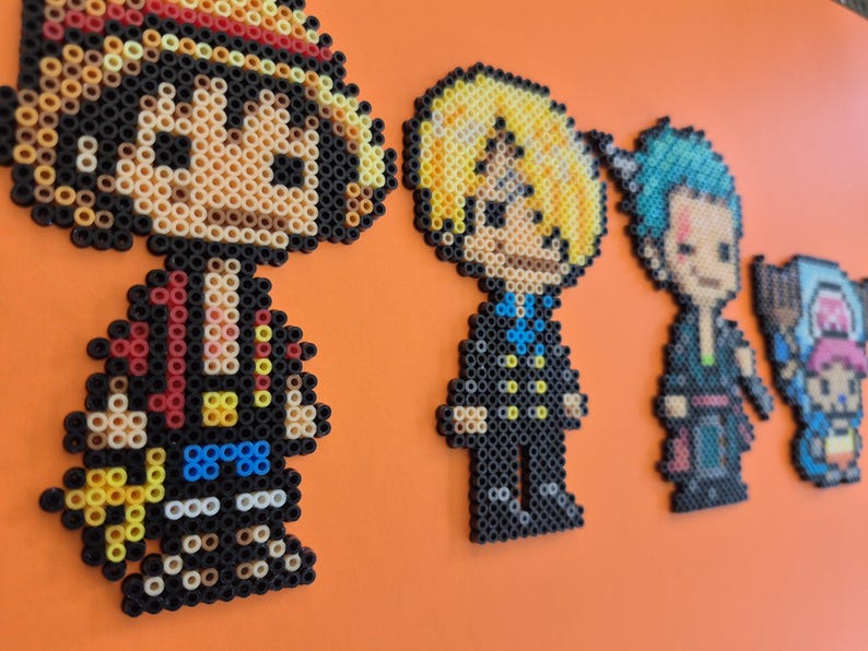 May include: Pixel art of four characters from the anime series One Piece. The characters are made from colorful beads and are arranged on an orange background. The first character is wearing a straw hat. The other characters are Sanji, Zoro, and Chopper.
