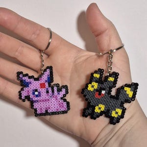 May include: Two pixel art keychains featuring Pokemon characters. The first keychain is a pink and purple Eevee with red eyes. The second keychain is a black and yellow Umbreon with red eyes.