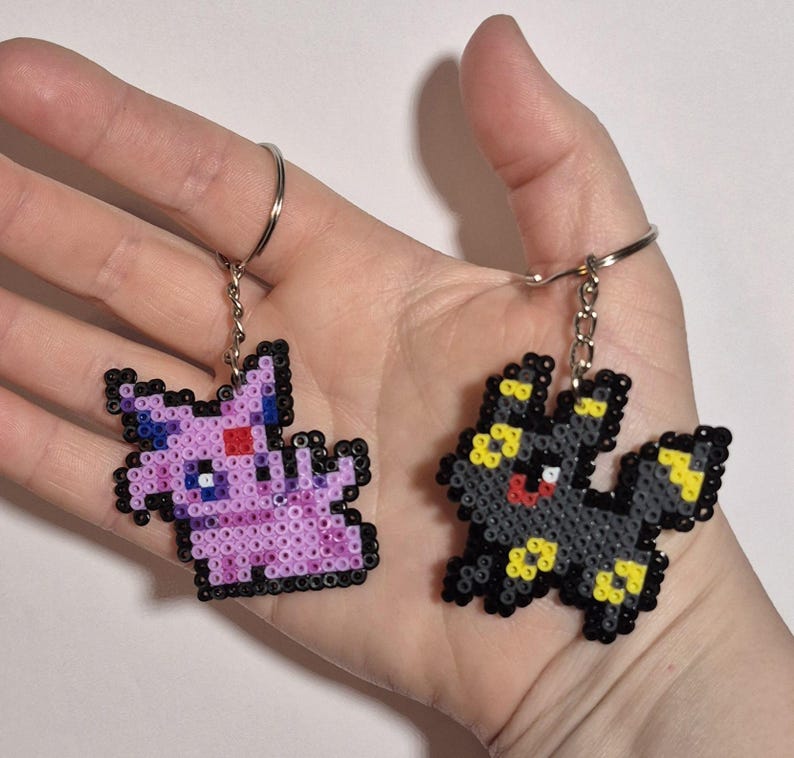 May include: Two pixel art keychains featuring Pokemon characters. The first keychain is a pink and purple Eevee with red eyes. The second keychain is a black and yellow Umbreon with red eyes.