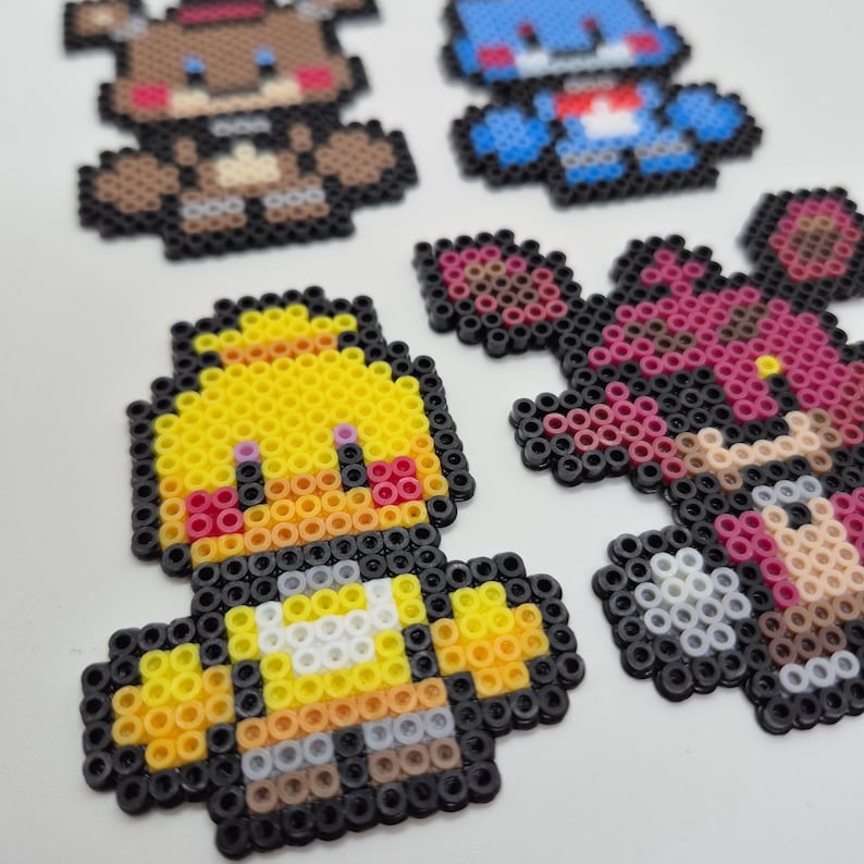 May include: Pixel art figures of characters in brown, blue, yellow, and pink. The figures are made of small, colorful beads, and are arranged on a white surface. The characters are in the shape of animals.