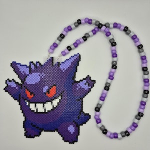 May include: A necklace featuring a purple pixelated character pendant made of fused plastic beads. The character has spiky ears, a wide grin with white teeth, and red eyes. The necklace strand consists of alternating purple, gray, and black beads.