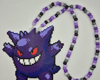 May include: A necklace featuring a purple pixelated character pendant made of fused plastic beads. The character has spiky ears, a wide grin with white teeth, and red eyes. The necklace strand consists of alternating purple, gray, and black beads.