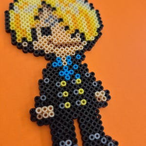 May include: A pixel art figure made from colorful plastic beads. The figure has a yellow head, a black outfit with blue and yellow accents, and is posed standing upright against an orange background.