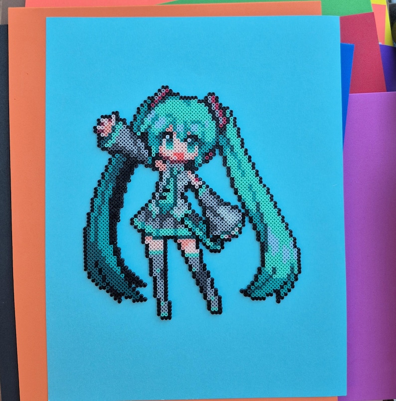 May include: A pixel art depiction of a character with long, turquoise pigtails, a matching outfit, and large eyes. The artwork is created with small, colorful beads on a light blue background, with orange and other colored paper in the background.