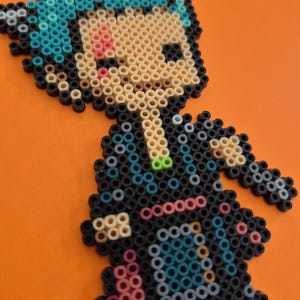 May include: A pixel art figure made from colorful plastic beads. The figure has blue hair, a black outfit, and a light skin tone. The background is a solid orange color.