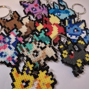 May include: A collection of colorful pixelated keychains featuring various Pokemon characters. The keychains are made of small plastic beads and are attached to silver key rings.