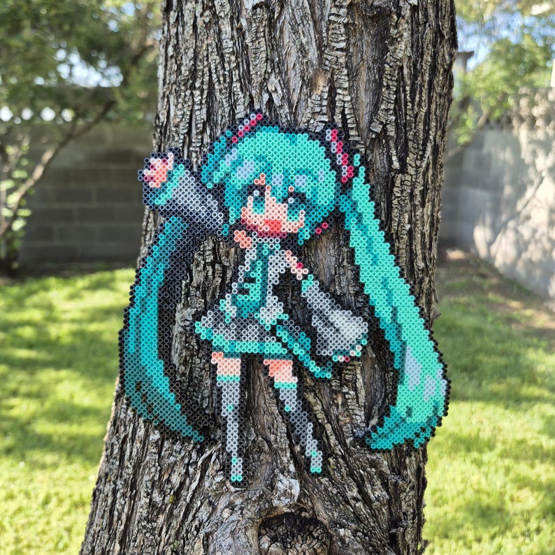 May include: A pixel art figure of a character with long, turquoise pigtails, a teal outfit, and red accents. The figure is made of small, colorful beads and is attached to a tree trunk. The background shows green grass and a blurred view of a building.