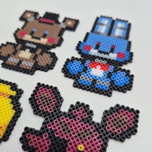 May include: A collection of colorful pixel art figures, including a brown bear with a black top hat, a blue car, and other characters. The figures are made from small, round beads, and are arranged on a white surface.