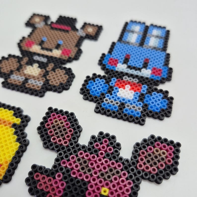 May include: A collection of colorful pixel art figures, including a brown bear with a black top hat, a blue car, and other characters. The figures are made from small, round beads, and are arranged on a white surface.