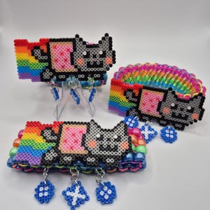May include: Three bracelets made of colorful plastic pony beads and pixelated perler bead charms. Each charm depicts a gray cat with a pink rectangular body and a rainbow trail. Small blue star-shaped beads hang from the bracelets by metal rings.