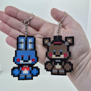 May include: Two keychains featuring pixelated characters. One is blue with a red bow tie, the other is brown with a black hat and red cheeks. Both have silver key rings attached, perfect for fans of video games.