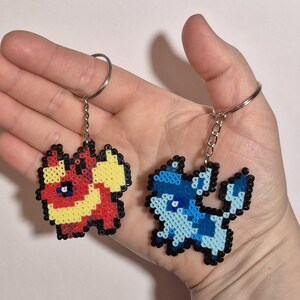 May include: Two pixel art keychains featuring Pokemon characters. The first keychain is red, yellow, and black and depicts a fire-type Pokemon. The second keychain is blue and white and depicts a water-type Pokemon.