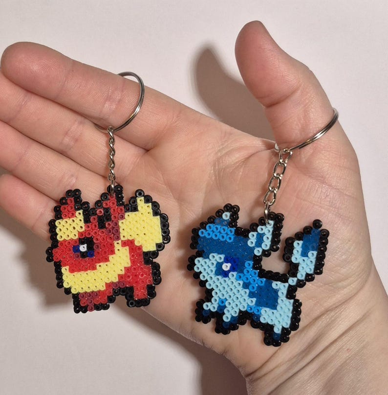 May include: Two pixel art keychains featuring Pokemon characters. The first keychain is red, yellow, and black and depicts a fire-type Pokemon. The second keychain is blue and white and depicts a water-type Pokemon.