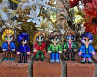 May include: Six pixel art figures made of colorful beads, standing on a brick surface. The figures are all different colors and have different hairstyles and clothing.