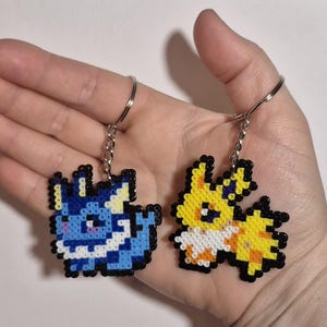 May include: Two pixel art keychains, one blue and one yellow, depicting cartoon characters. The blue keychain features a blue and white character with a white collar and yellow ears. The yellow keychain features a yellow and white character with a white collar and yellow ears.
